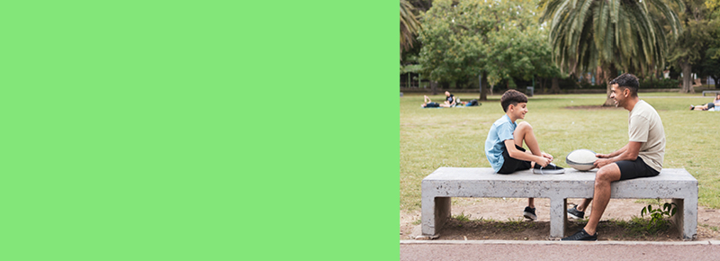 A man and a boy sit on a concrete bench in a park, holding a frisbee. The background is a grassy area with trees.