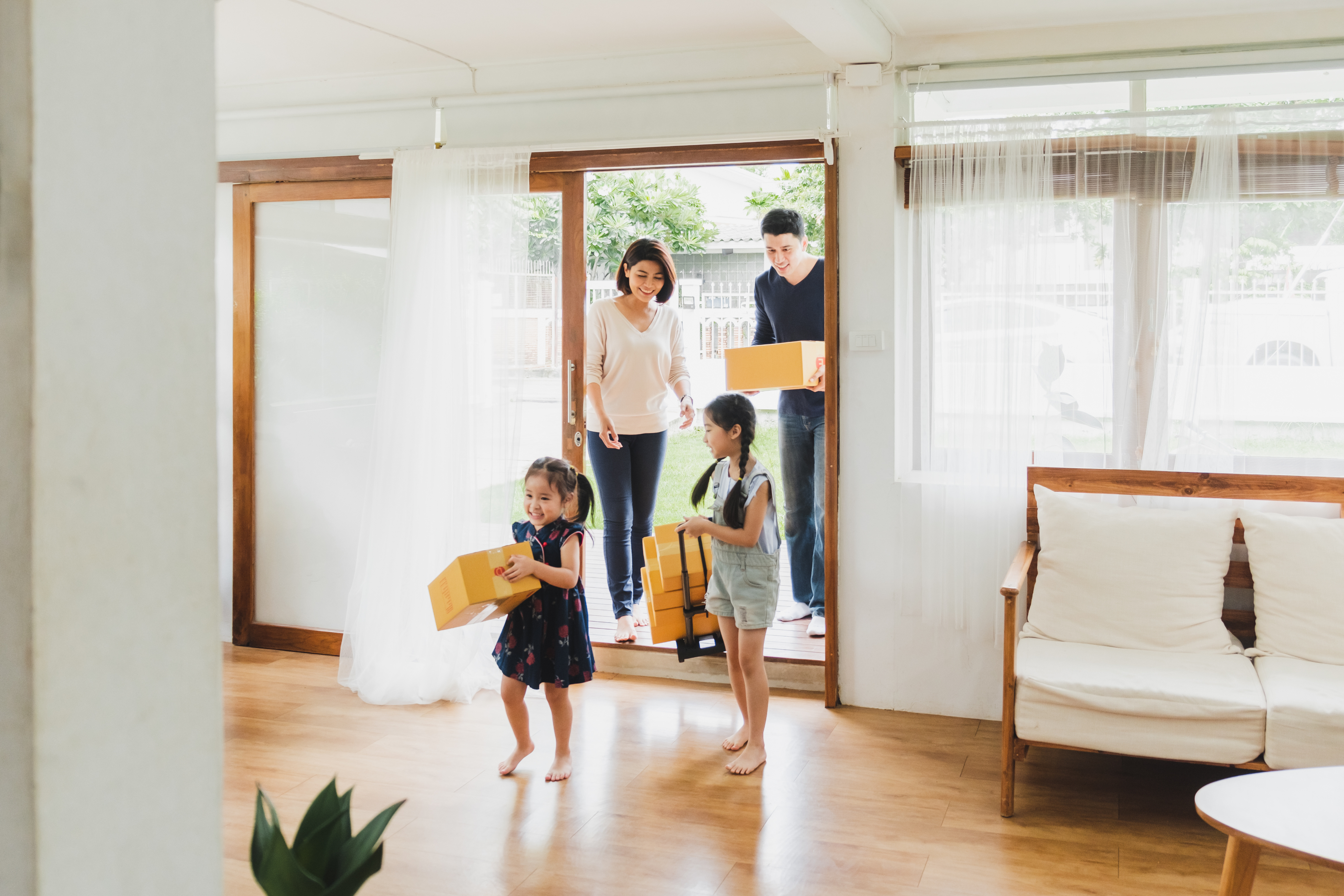 Family with two children carrying boxes enters a sunlit living room through sliding glass doors.