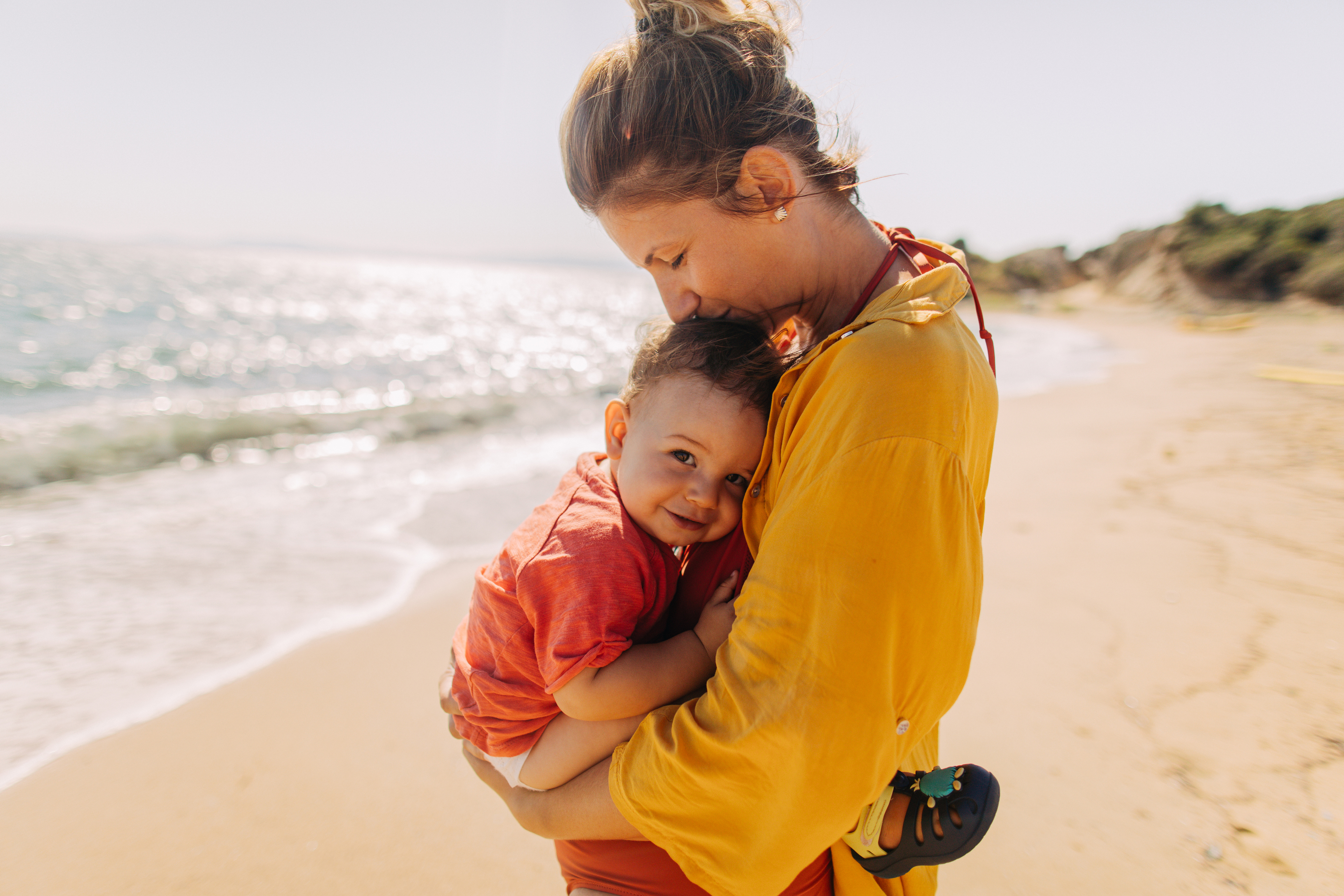 A woman embraces a child on the sandy beach, surrounded by the ocean's waves and a bright, sunny sky.