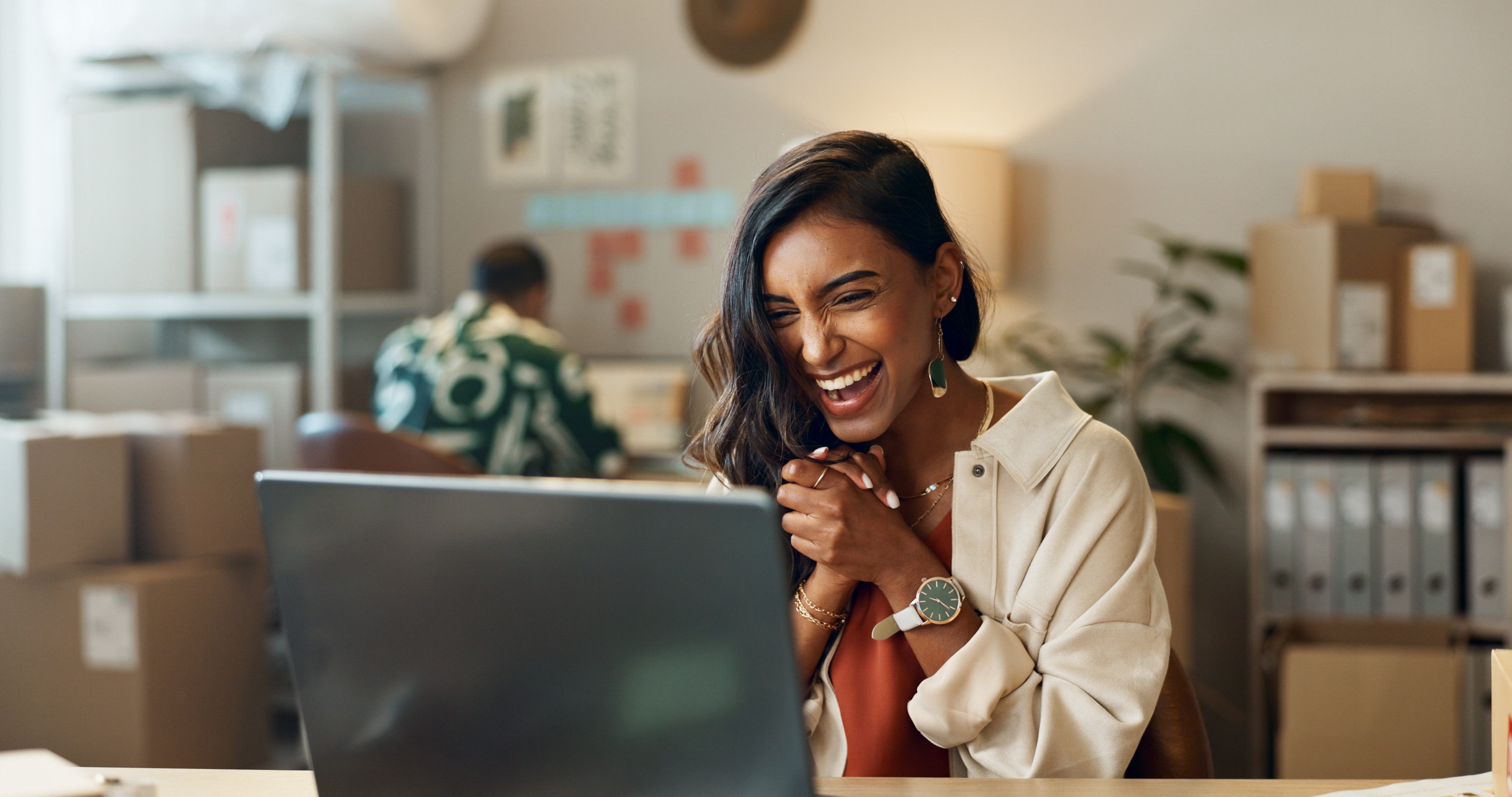 A woman smiles while seated at a desk, working on her laptop, exuding a sense of positivity and focus.