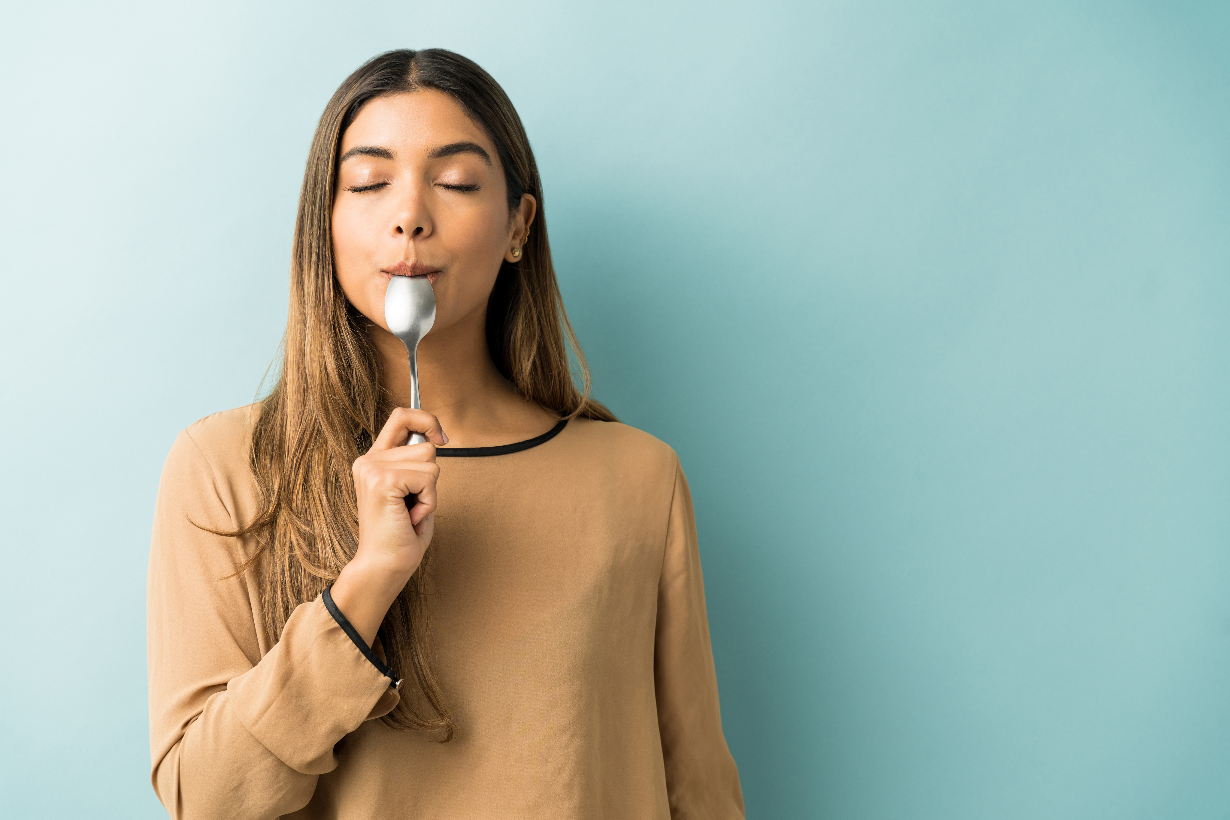 Women with spoon in mouth, eyes closed,  looking happy and satisfied against a blue wall.